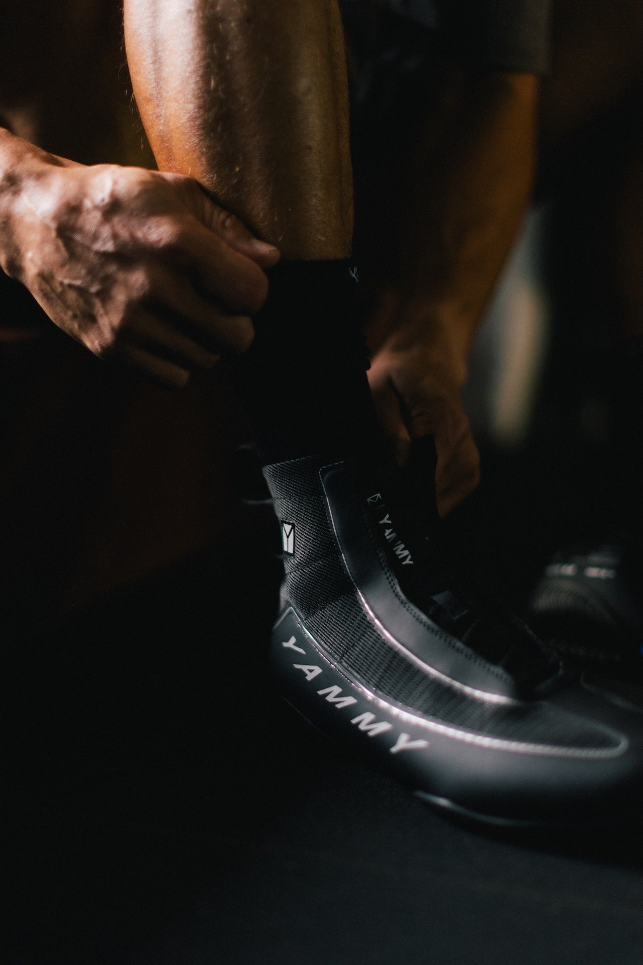 A close-up lifestyle shot of a cyclist putting on a black cycling shoe with 'YAMMY' branding, with their legs and hands visible in a dark, moody setting.