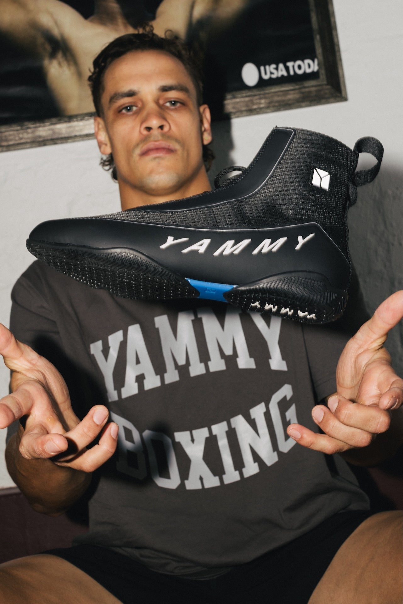 A man wearing a black YAMMY boxing shirt holds up a black YAMMY boxing shoe in front of his face. The shoe features white 'YAMMY' branding on the side and a small white logo on the ankle collar, with a small blue accent on the midsole.