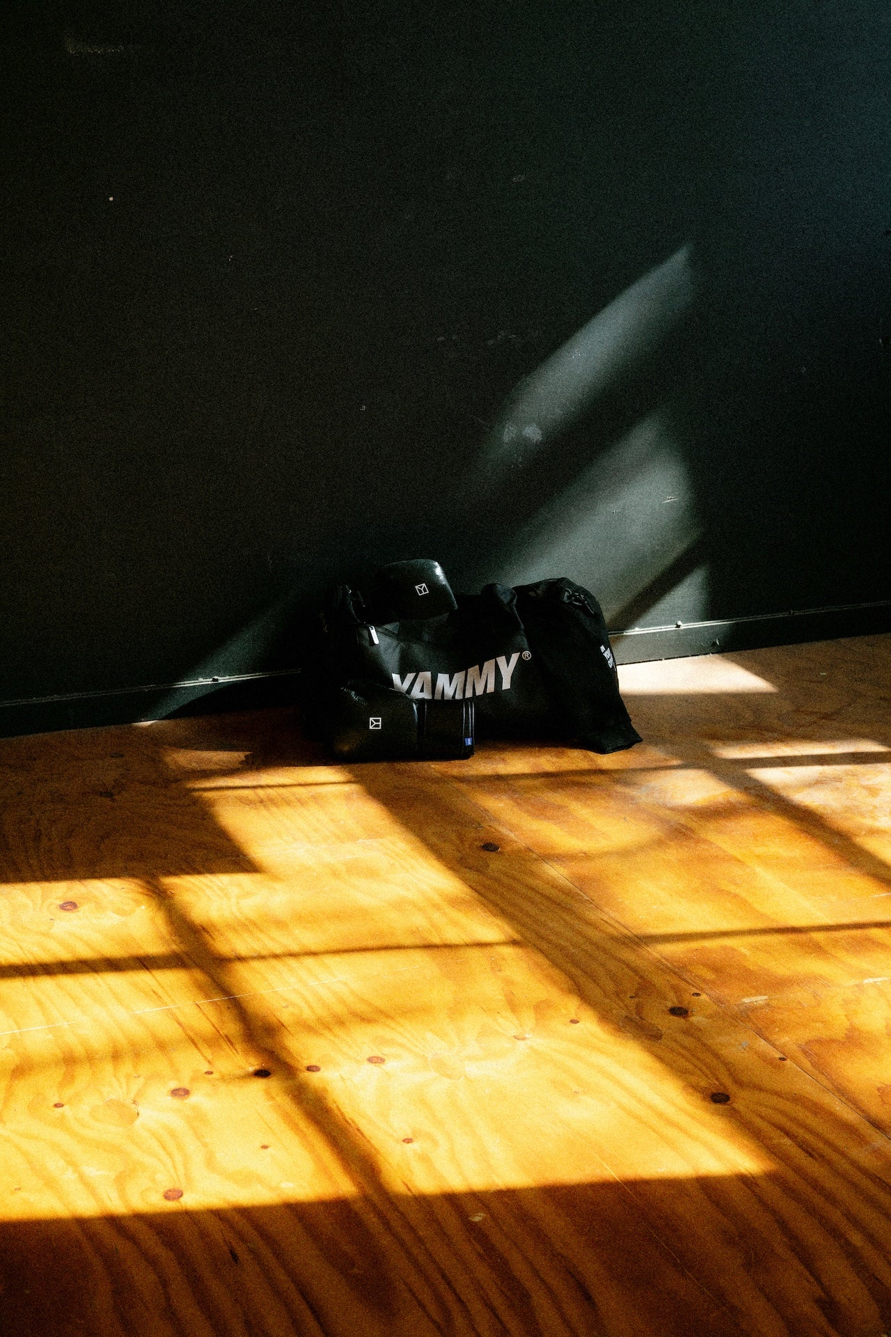 A black duffel bag with the word 'YAMMY' printed in white across the front, resting on a wooden floor in dramatic sunlight and shadow against a dark wall.