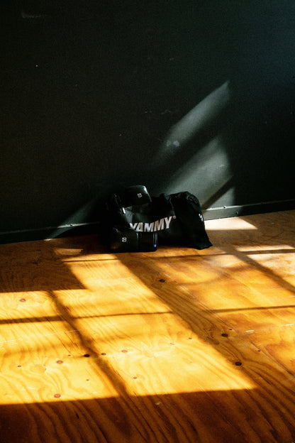 A black duffel bag with the word 'YAMMY' printed in white across the front, resting on a wooden floor in dramatic sunlight and shadow against a dark wall.