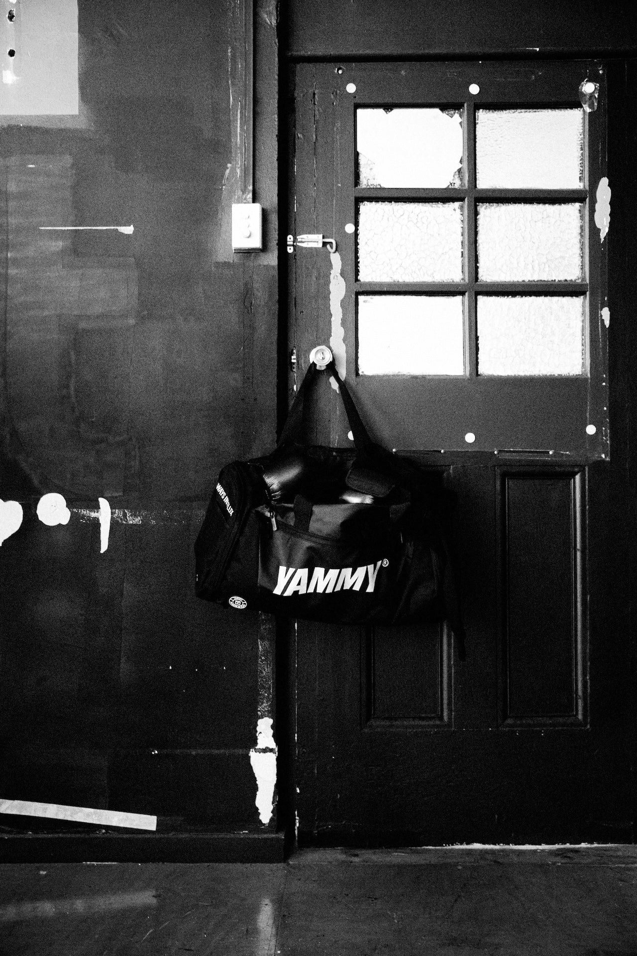 A black duffel bag with the word 'YAMMY' printed in white across the front, hanging from a door knob on a dark paneled door in a black-and-white photo.