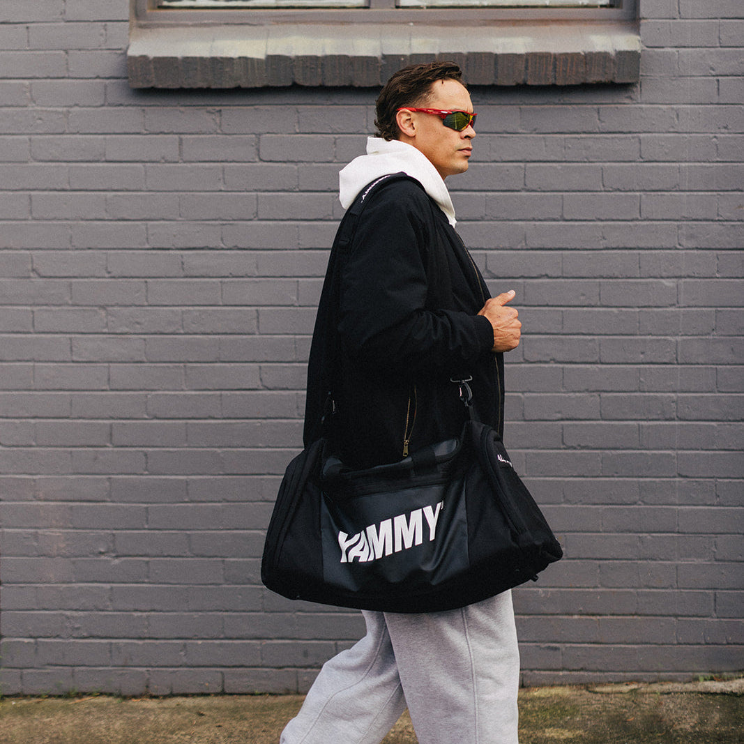 A man wearing sunglasses and a black jacket walks past a gray brick building  carrying a black duffel bag.