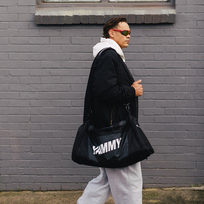 A man wearing sunglasses and a black jacket walks past a gray brick building  carrying a black duffel bag.