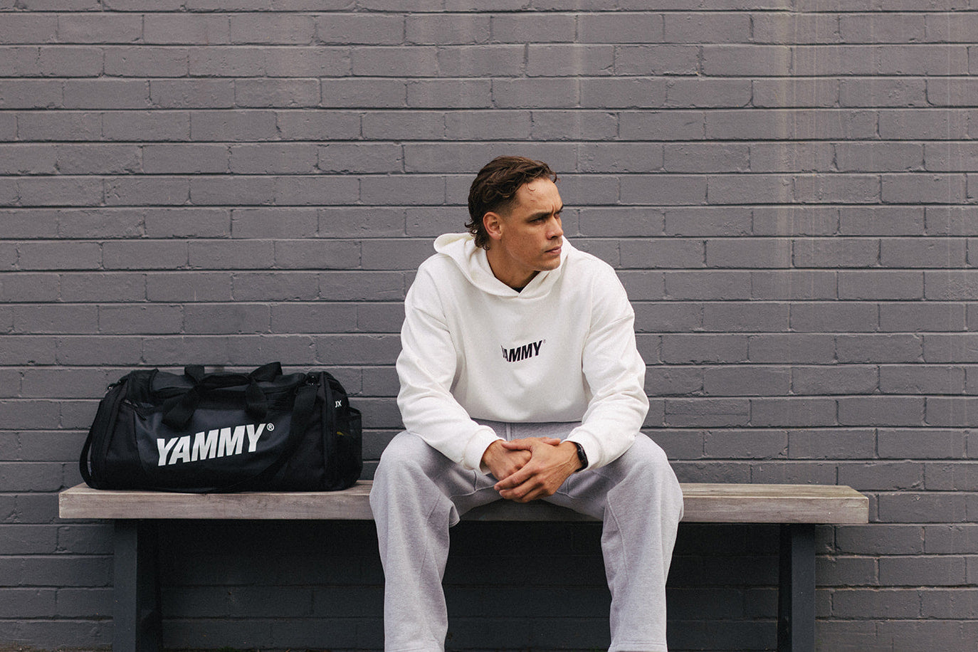 A man sits on a bench against a gray brick wall, wearing a white Yammy hoodie and sweatpants with blue sneakers. A black Yammy duffel bag rests on the bench beside him.