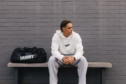 A man sits on a bench against a gray brick wall, wearing a white Yammy hoodie and sweatpants with blue sneakers. A black Yammy duffel bag rests on the bench beside him.