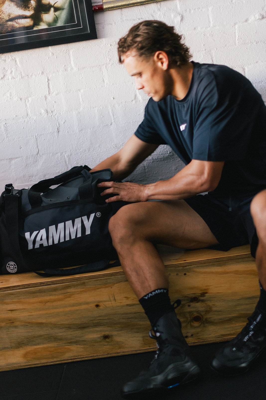 A man in a gym setting sits on a wooden bench next to a black Yammy duffel bag, wearing a dark navy athletic t-shirt and shorts.