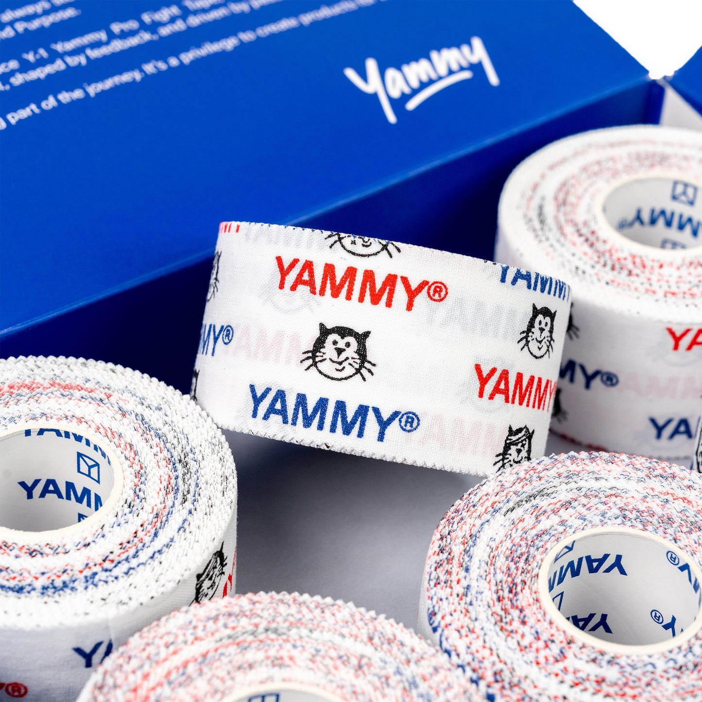 Several rolls of white athletic tape with red and blue speckled edges and repeated YAMMY logos and Hector cat icons, arranged in front of a blue branded box.