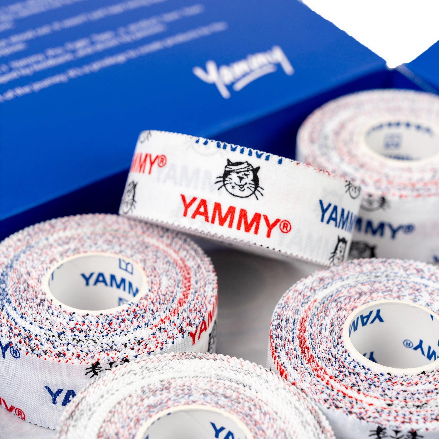 Several rolls of white elastic tape with red and blue speckled edges and printed YAMMY branding, arranged in front of a blue box.