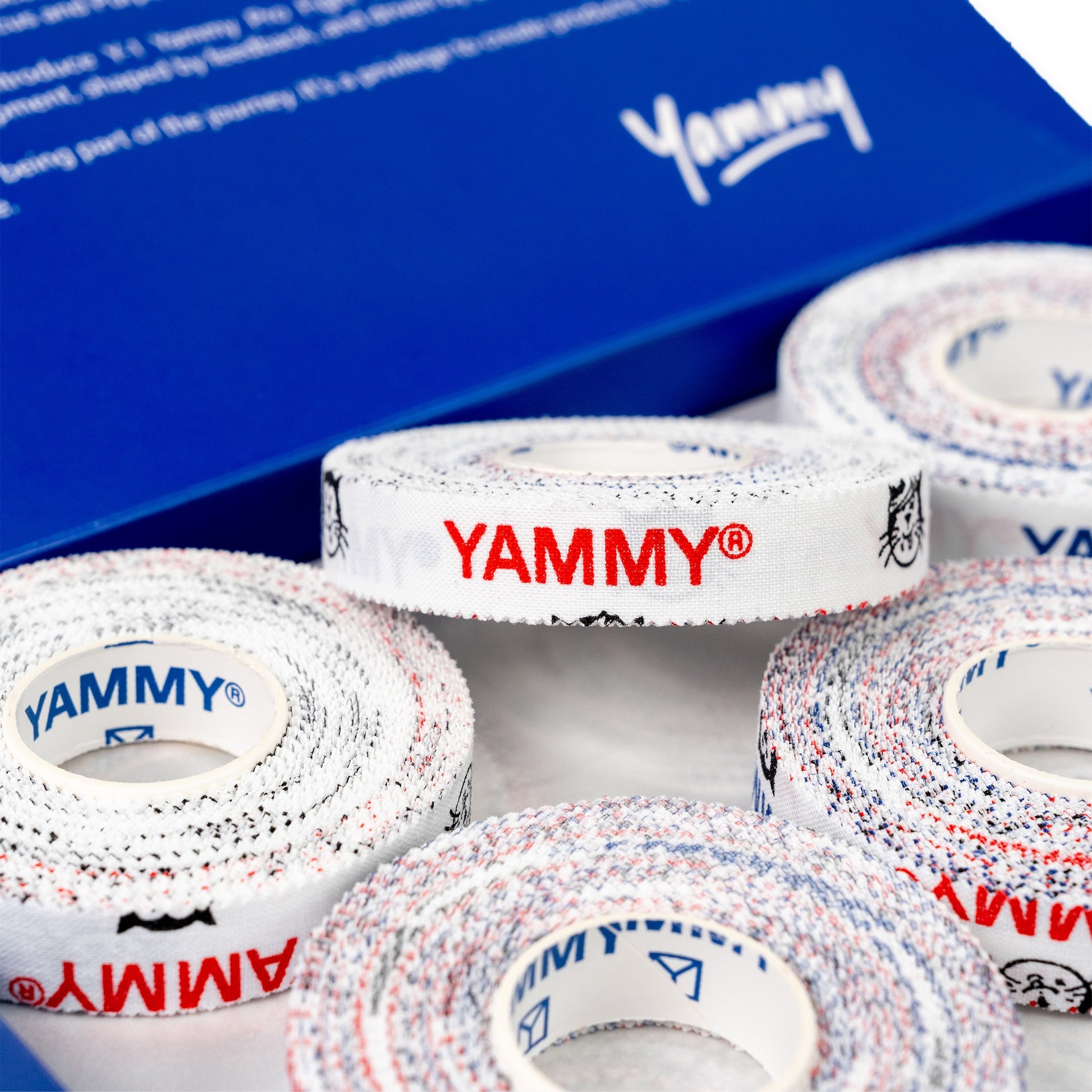 Several rolls of YAMMY brand double-sided tape are arranged in front of a blue branded box. The tape has a white base with red and blue speckled edges and red and blue YAMMY logos printed on the outer side.