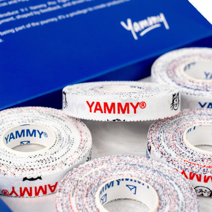 Several rolls of YAMMY brand double-sided tape are arranged in front of a blue branded box. The tape has a white base with red and blue speckled edges and red and blue YAMMY logos printed on the outer side.