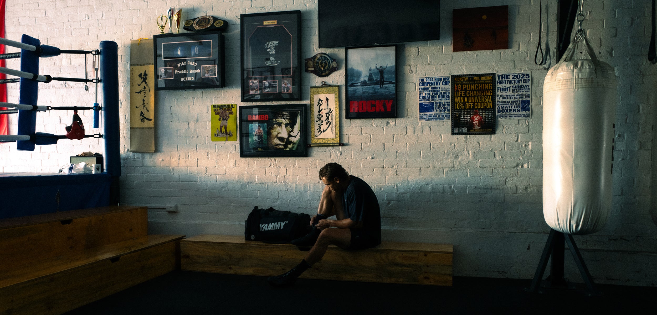 A person sits on a wooden bench in a dimly lit boxing gym, with a punching bag on the right, a boxing ring on the left, and framed posters and signs on a white brick wall behind them.