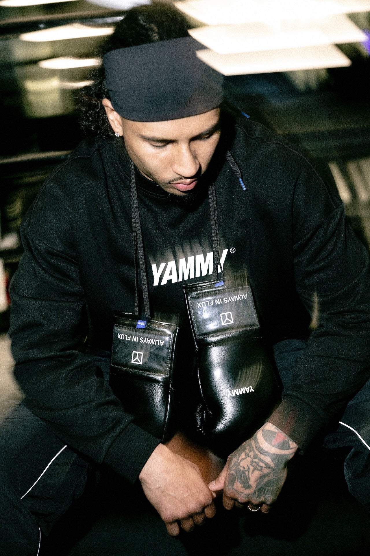 A man in a black Yammy gym hoodie and black headband sits on the floor, holding a pair of black Yamm boxing gloves. The Yamm logo is clearly visible on the hoodie and the gloves.
