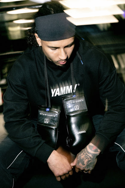 A man in a black Yammy gym hoodie and black headband sits on the floor, holding a pair of black Yamm boxing gloves. The Yamm logo is clearly visible on the hoodie and the gloves.