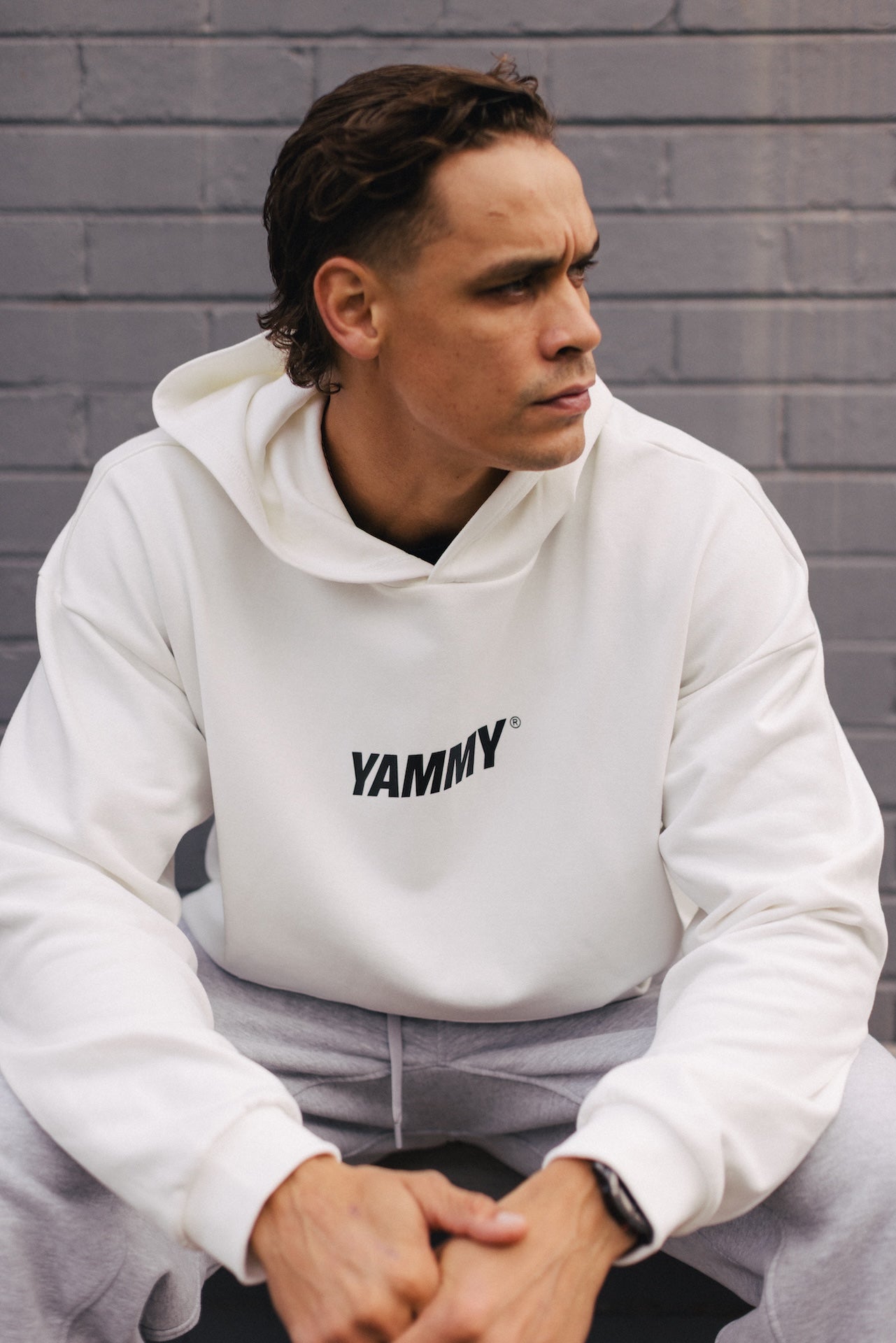 A man wearing a white pullover hoodie with 'YAMMY' printed in black across the chest, paired with light gray sweatpants, sitting against a gray brick wall.