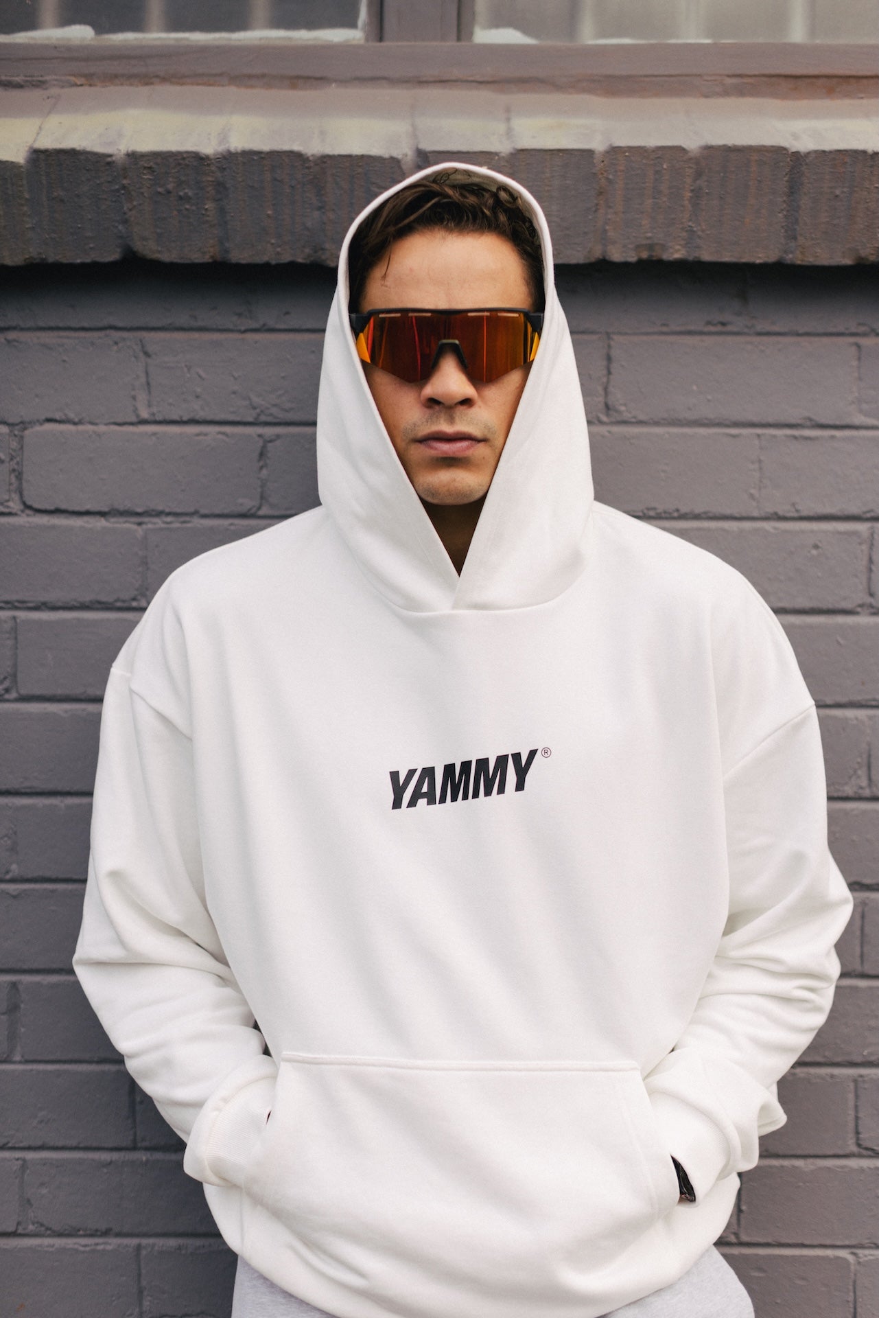 A man wearing a white pullover hoodie with 'YAMMY' printed in black across the chest, standing against a gray brick wall.