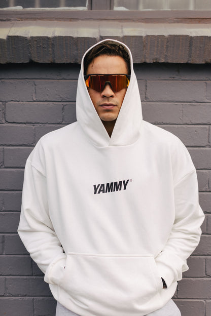 A man wearing a white pullover hoodie with 'YAMMY' printed in black across the chest, standing against a gray brick wall.