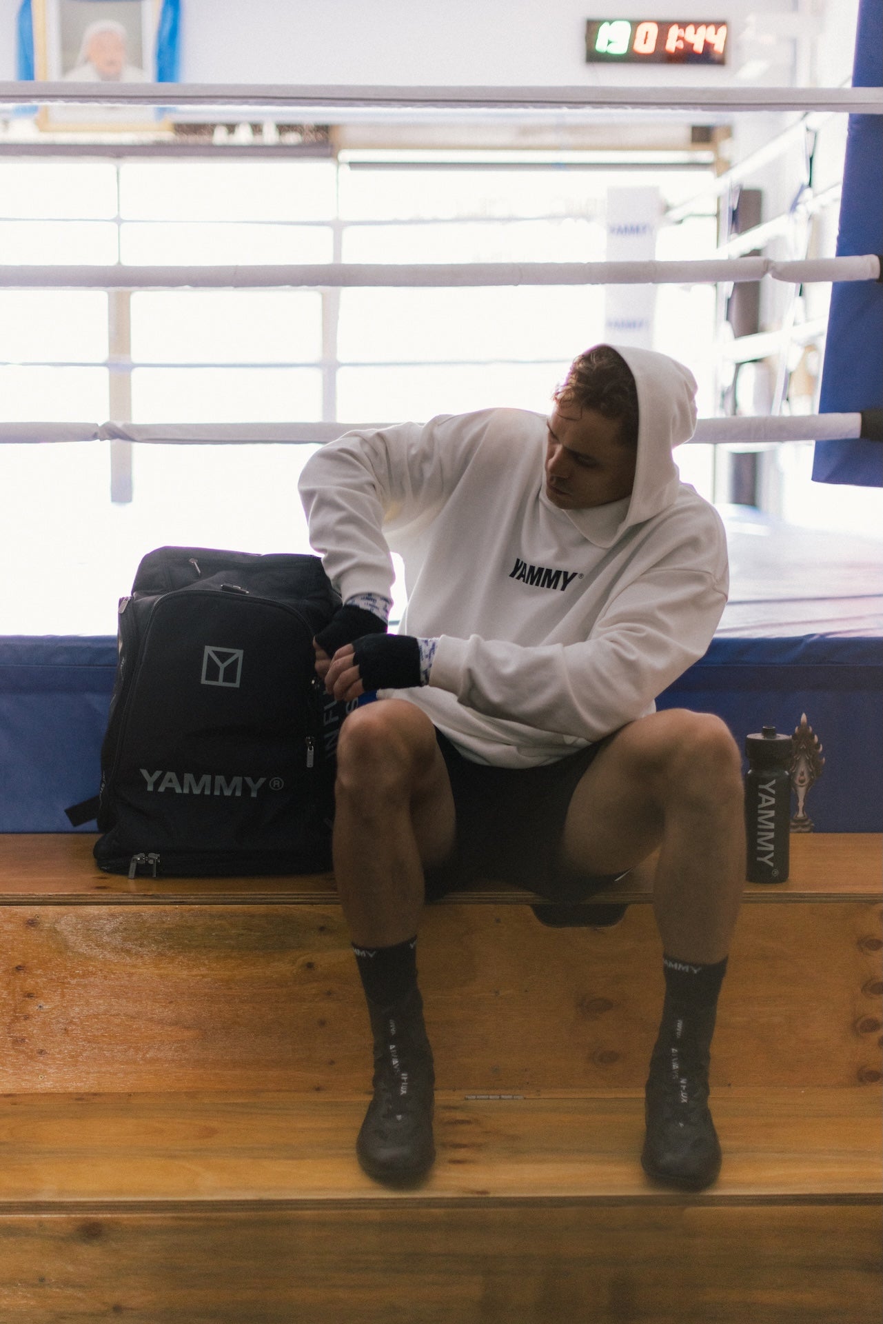 A man sits on wooden boxing steps inside a boxing ring, wearing a white YAMMY hoodie, black shorts, and black socks, with a black YAMMY bag beside him.