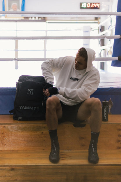 A man sits on wooden boxing steps inside a boxing ring, wearing a white YAMMY hoodie, black shorts, and black socks, with a black YAMMY bag beside him.