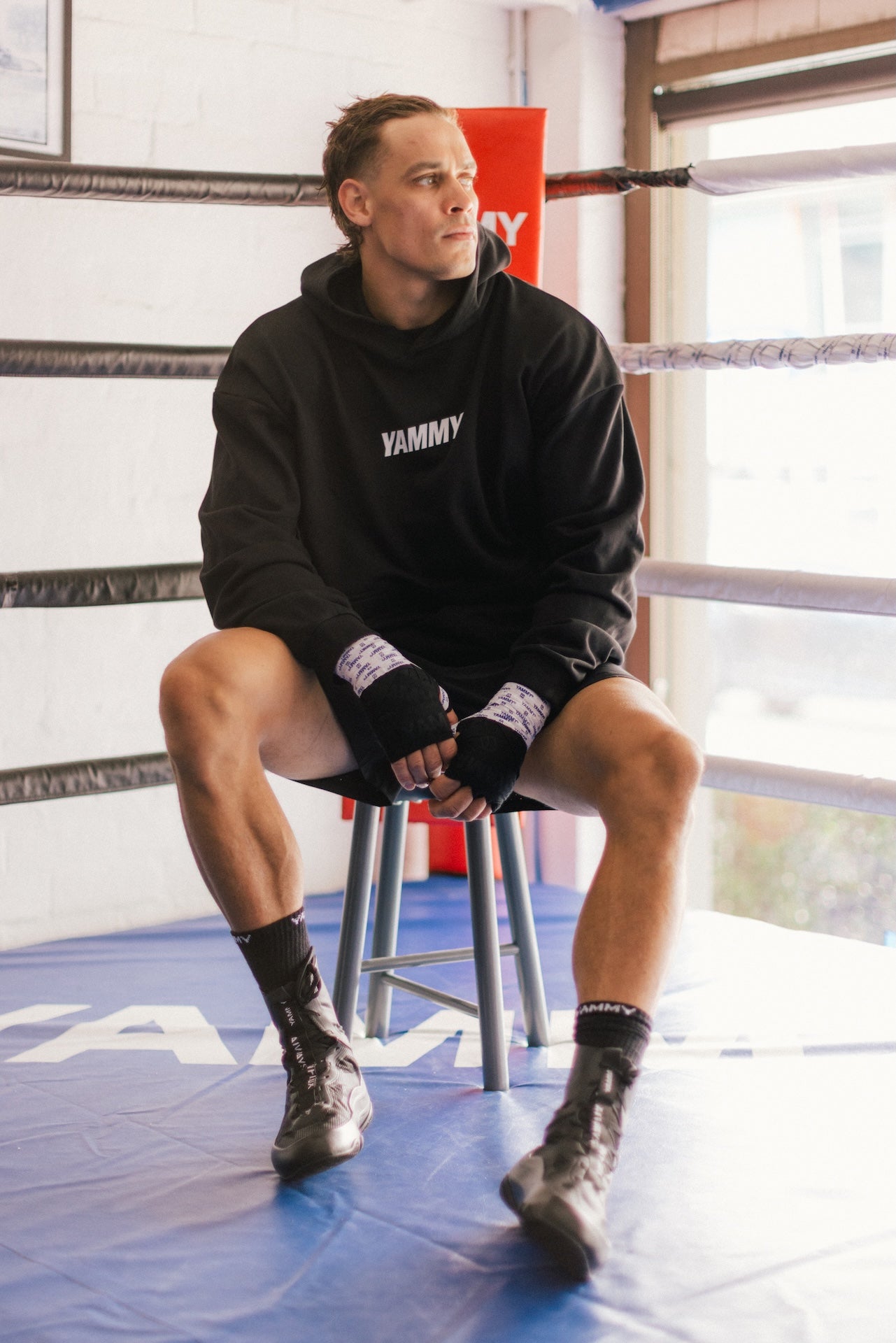A man sits on a metal stool inside a boxing ring wearing a black hoodie with 'YAMMY' printed in white on the chest, black boxing shorts, black socks, and boxing boots.