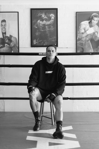 A man sits on a stool inside a boxing ring wearing a black hoodie with 'YAMMY' printed on the chest, boxing shorts, and boots. Three framed boxing portraits hang on the white brick wall behind him.