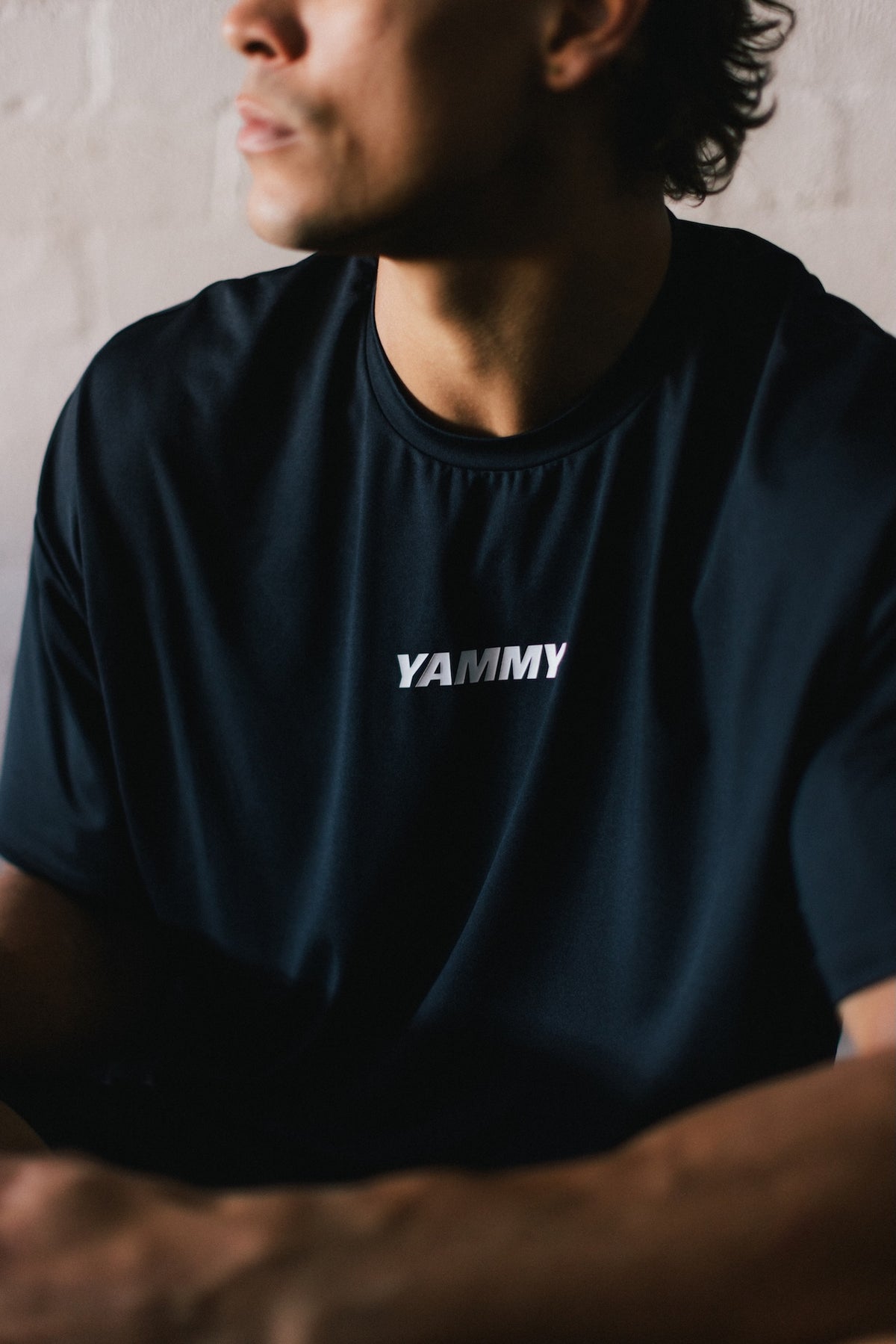 A close-up of a man wearing a black t-shirt with the word 'YAMMY' printed in white across the chest, set against a neutral light brick wall background.