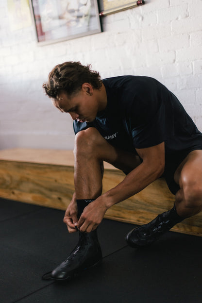 A man in a gym crouching down to tie his black athletic boot, wearing a dark navy t-shirt and black shorts in a room with a white brick wall and wooden bench.