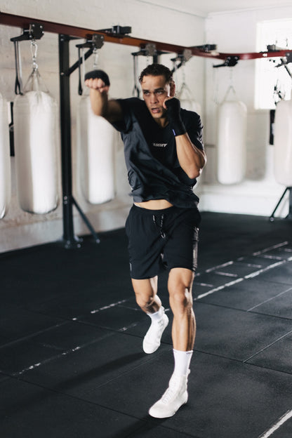 A man in a gym wearing a black athletic t-shirt, black shorts, white socks, and white boxing shoes, captured mid-motion as he throws a punch. Heavy bags hang in the background of the gym.