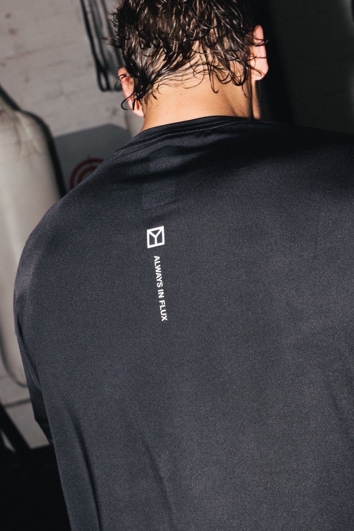 A close-up back view of a person wearing a black long-sleeve shirt with a small white vertical logo and text on the upper back, reading 'ALWAYS IN FLUX' with a geometric symbol above it.