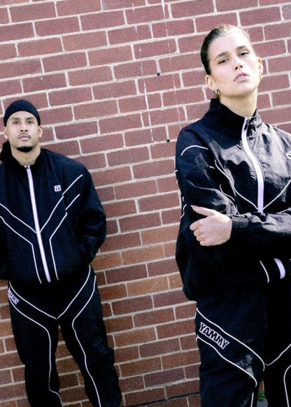 Two models wearing matching black tracksuits with white piping details, standing in front of a red brick wall on an asphalt surface.