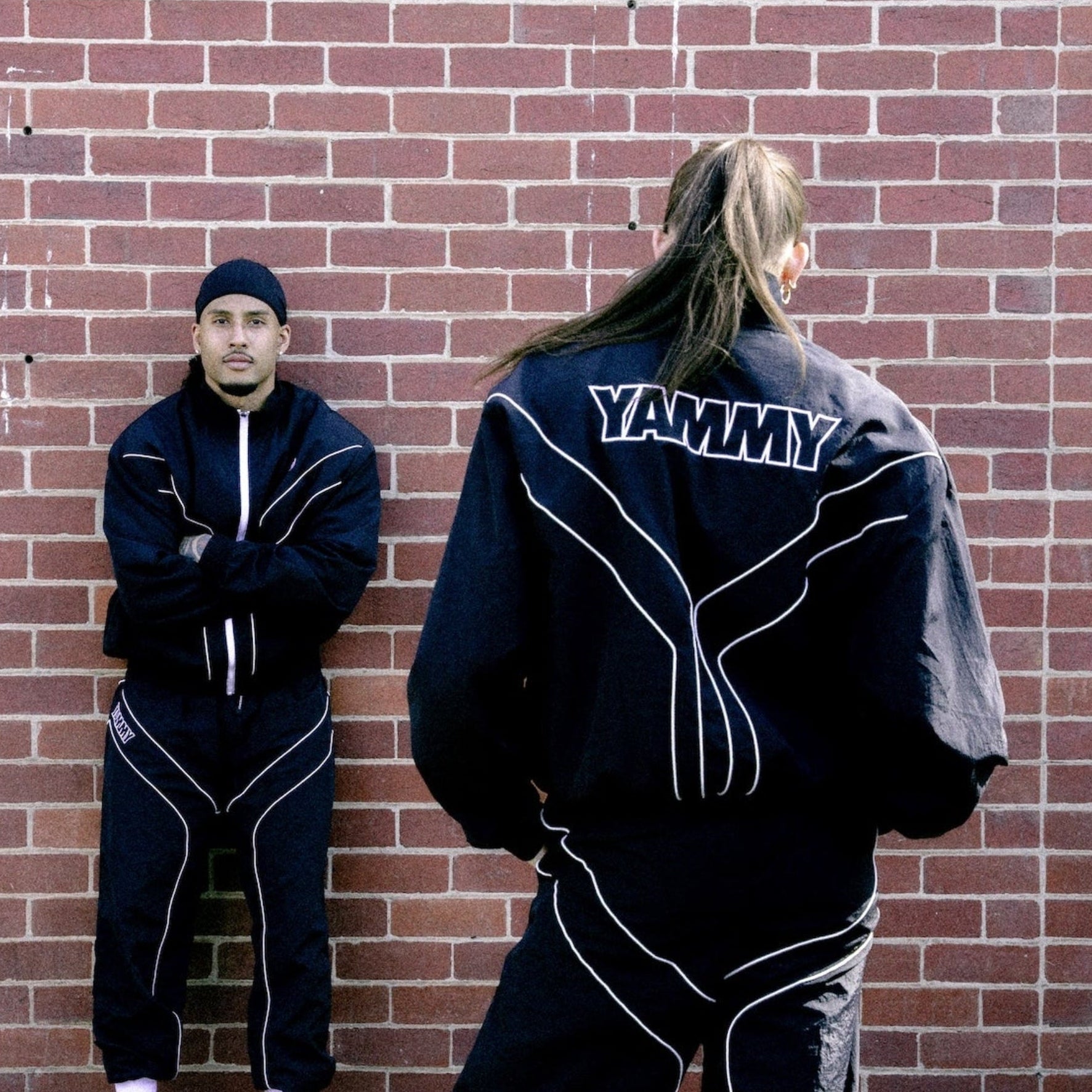 Two people wearing matching black tracksuits with white piping details, standing in front of a red brick wall. One person is facing forward showing the front of the jacket with 'YAMMY' printed on the back, and the other is facing sideways showing the side of the tracksuit.