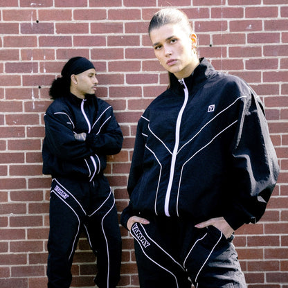 Two models wearing matching black tracksuits with white piping details, standing in front of a red brick wall.