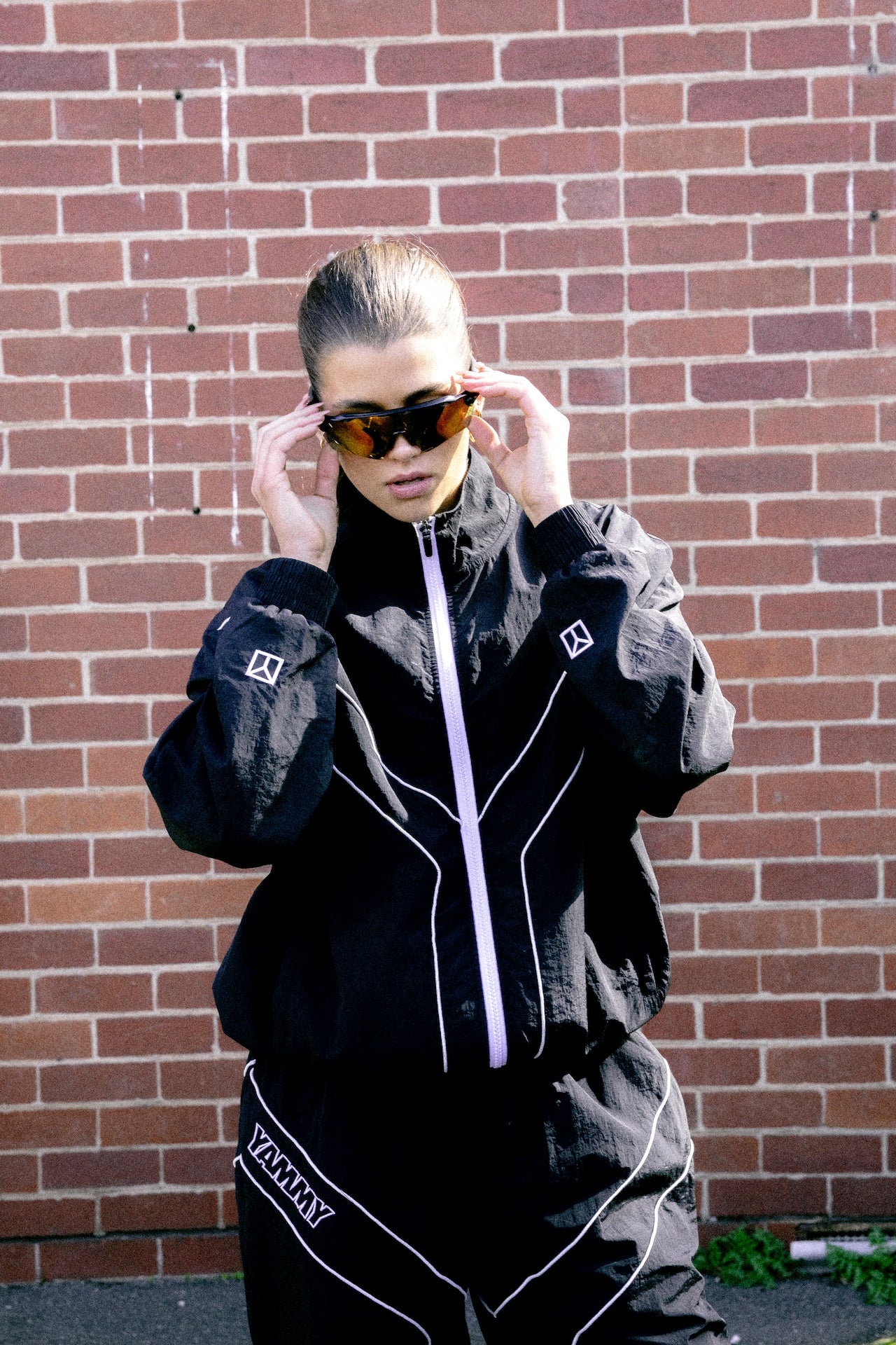 A female model wearing a black zip-up jacket and matching black pants with white piping and 'YAMMY' text, adjusting dark sunglasses in front of a red brick wall.