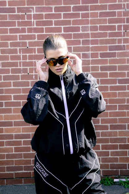 A female model wearing a black zip-up jacket and matching black pants with white piping and 'YAMMY' text, adjusting dark sunglasses in front of a red brick wall.