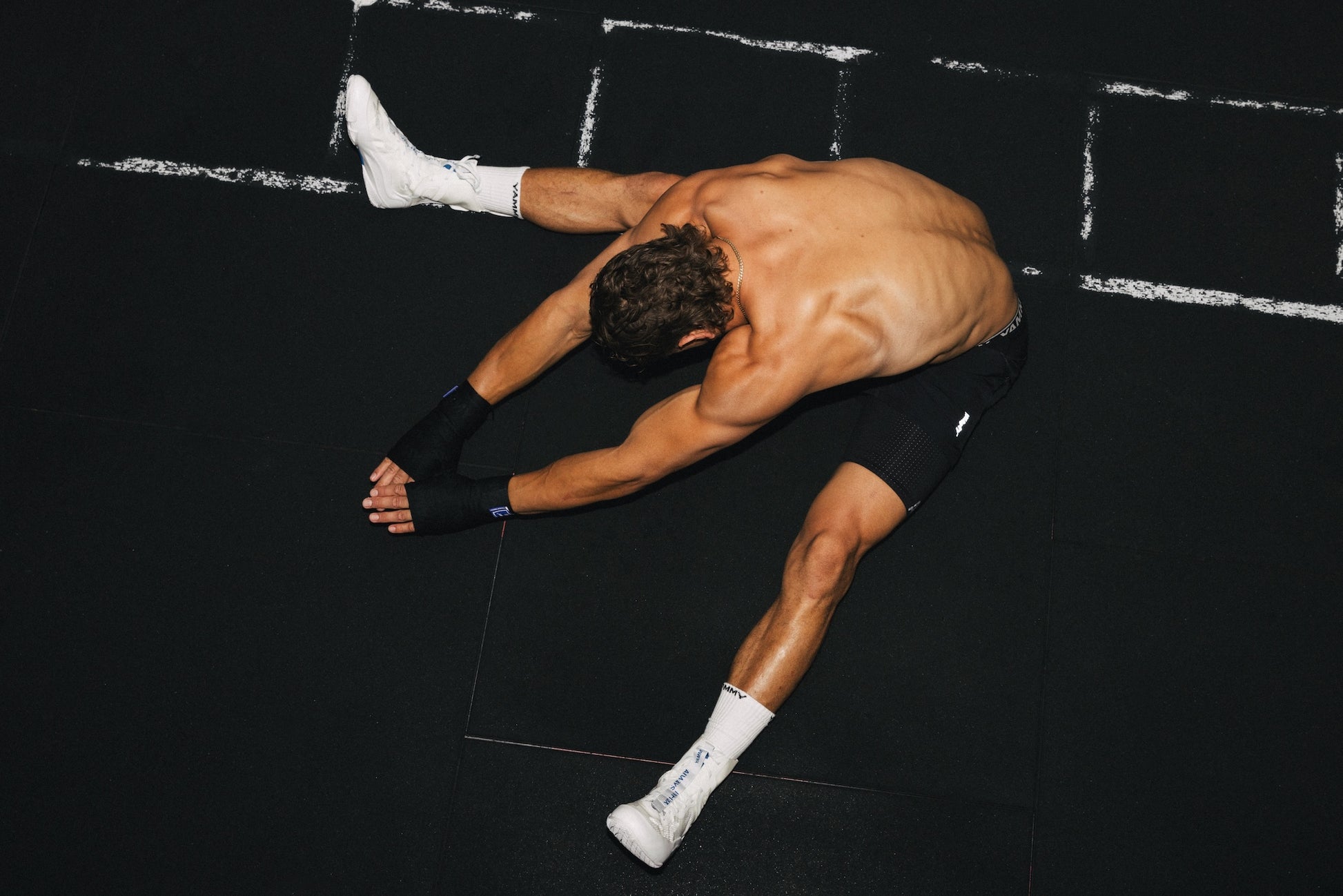 A shirtless man in black athletic shorts and white socks stretches forward on a black gym floor.