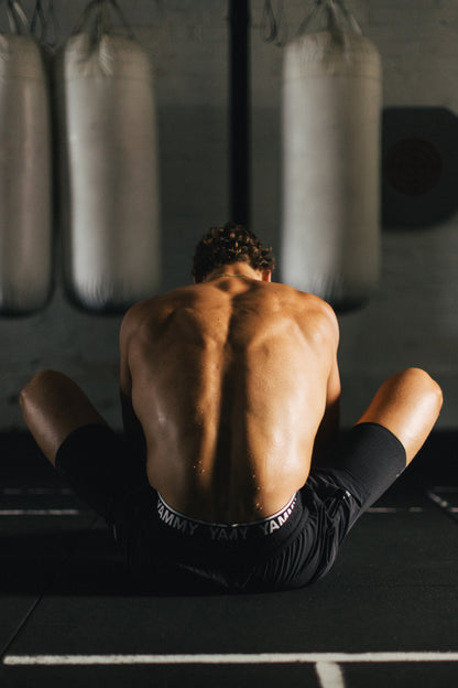 A shirtless man sits on his knees on a gym floor with his back to the camera, wearing black athletic shorts with a visible YAMMY Boxing waistband. Heavy bags hang in the background of a dimly lit gym.