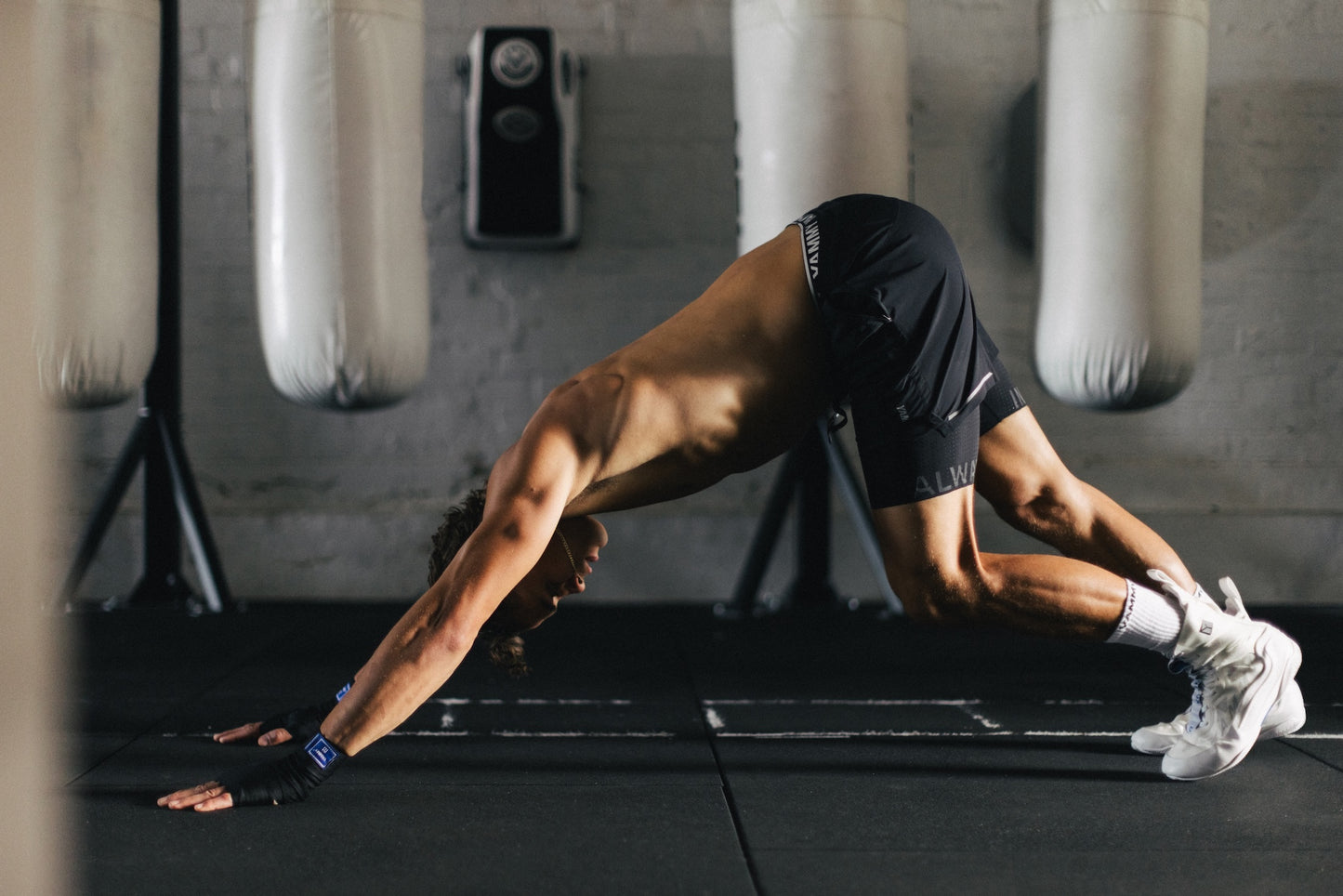 A shirtless man in black athletic shorts and white sneakers performs a yoga stretch on a black gym floor, with hanging punching bags and a gray brick wall in the background.