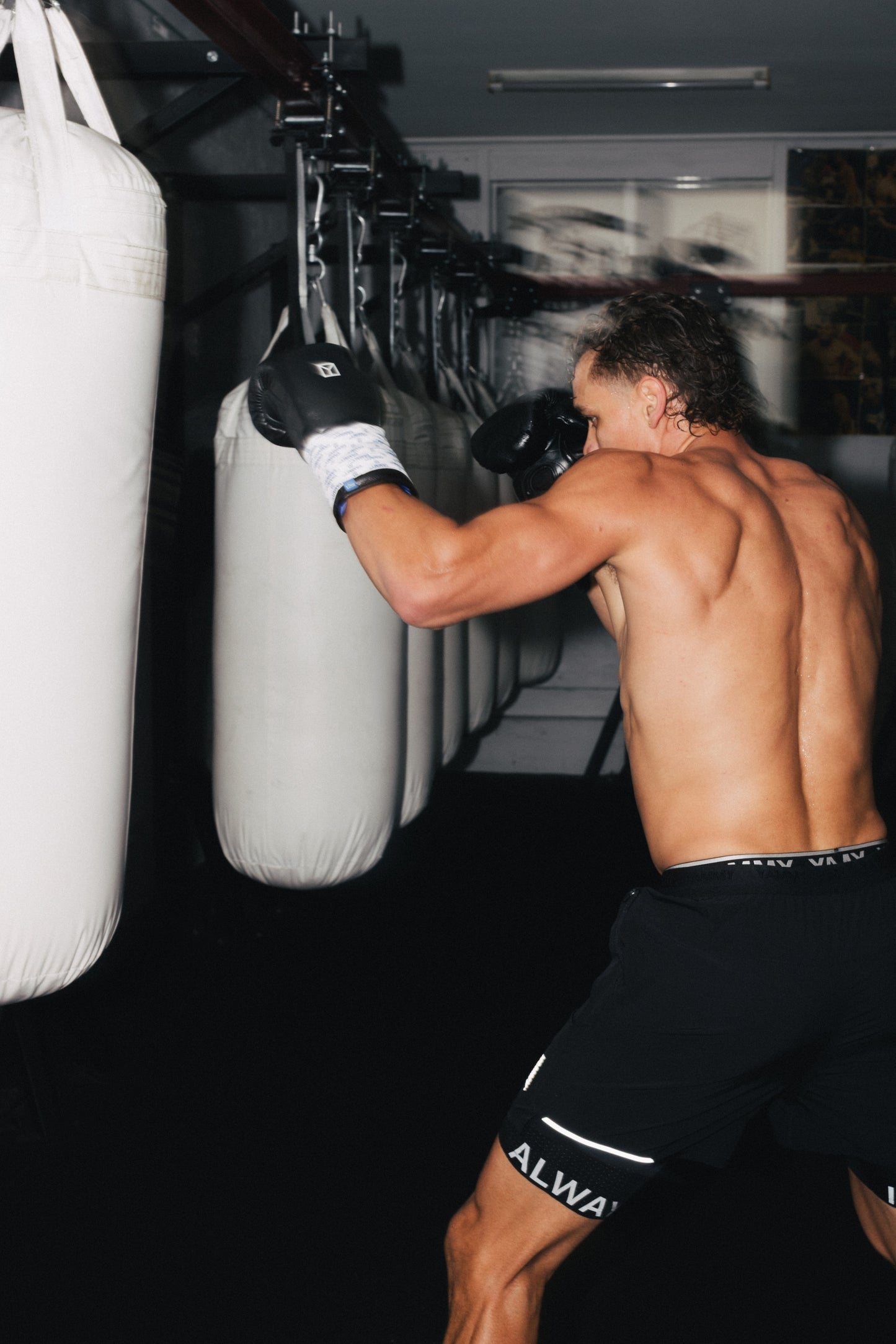 A shirtless man wearing black boxing gloves and black athletic shorts punches a white hanging punching bag in a dark gym.