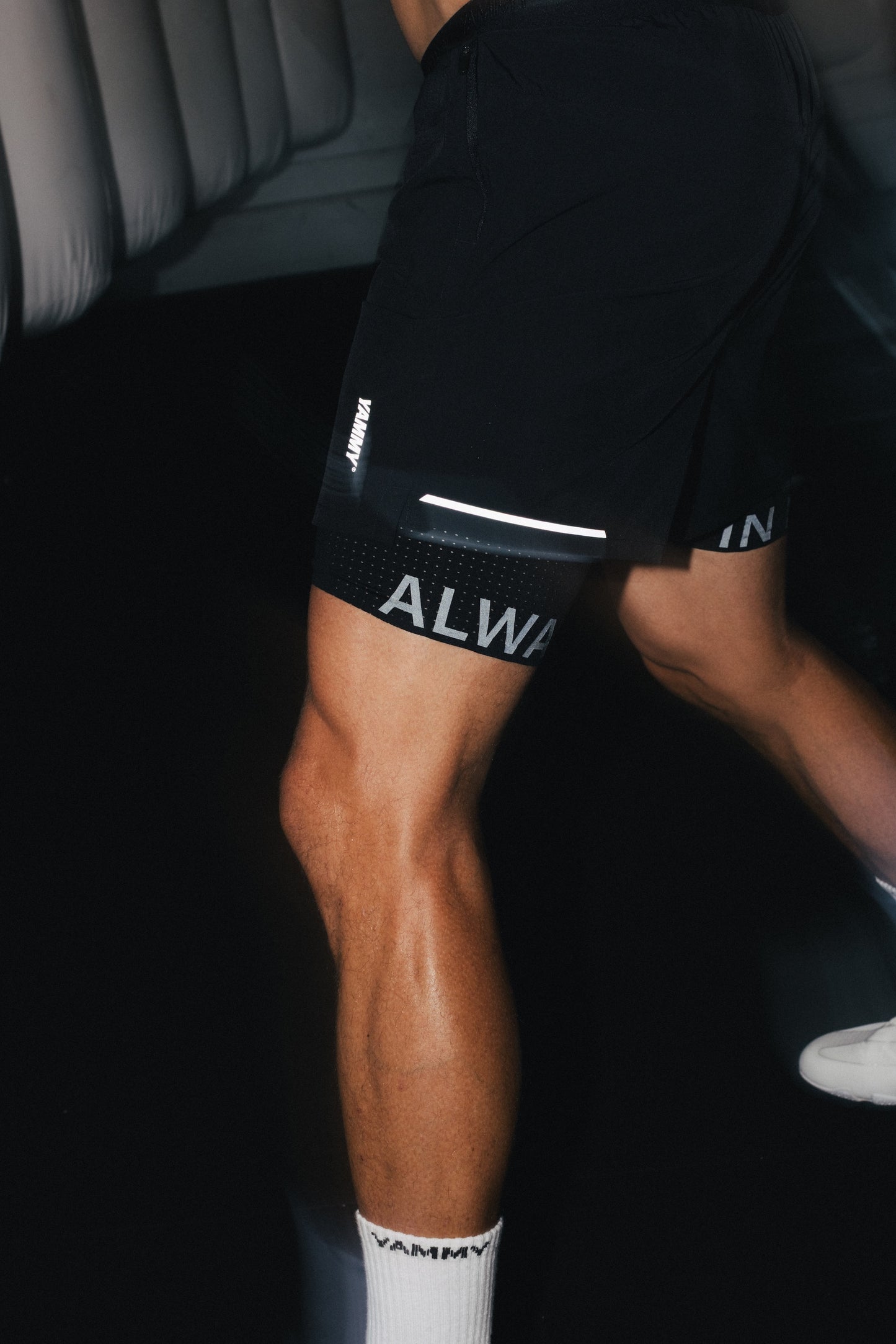 A close-up lifestyle shot of a person's leg wearing black athletic shorts with white branding text and a white reflective stripe, paired with white socks.