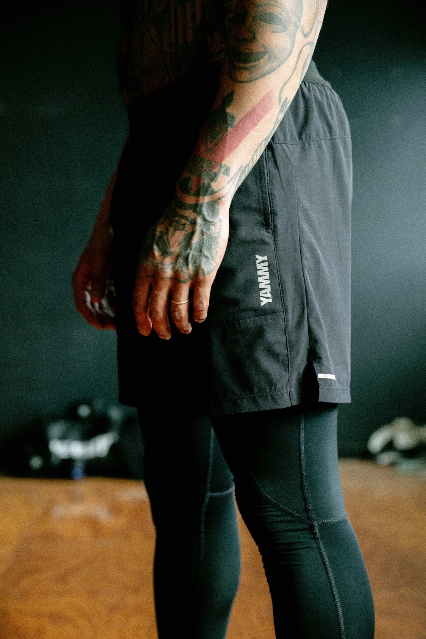 A close-up lifestyle shot of a person wearing black athletic shorts with a white vertical 'YAMMY' logo and black leggings, standing in a dimly lit gym environment.