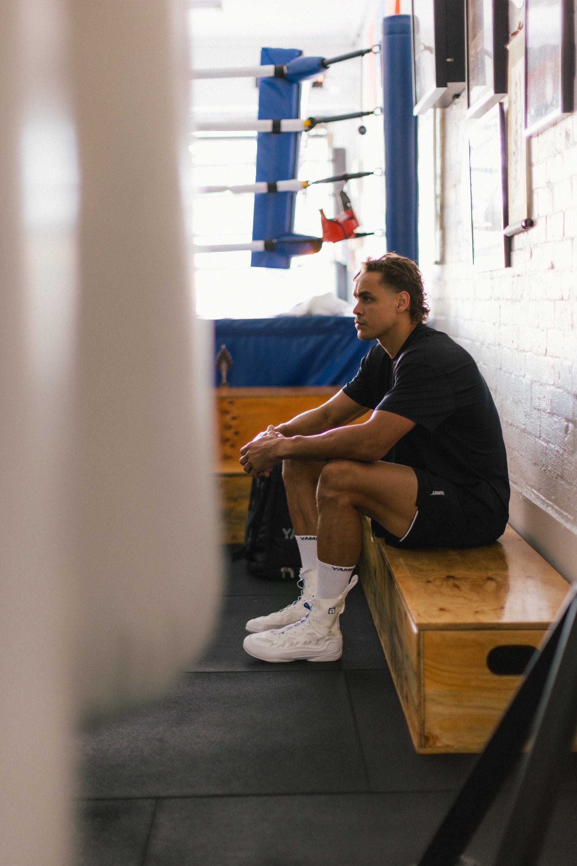 A man sits on a wooden bench in a gym, wearing a black t-shirt, black shorts, white socks, and white boxing shoes. A boxing ring is visible in the background.