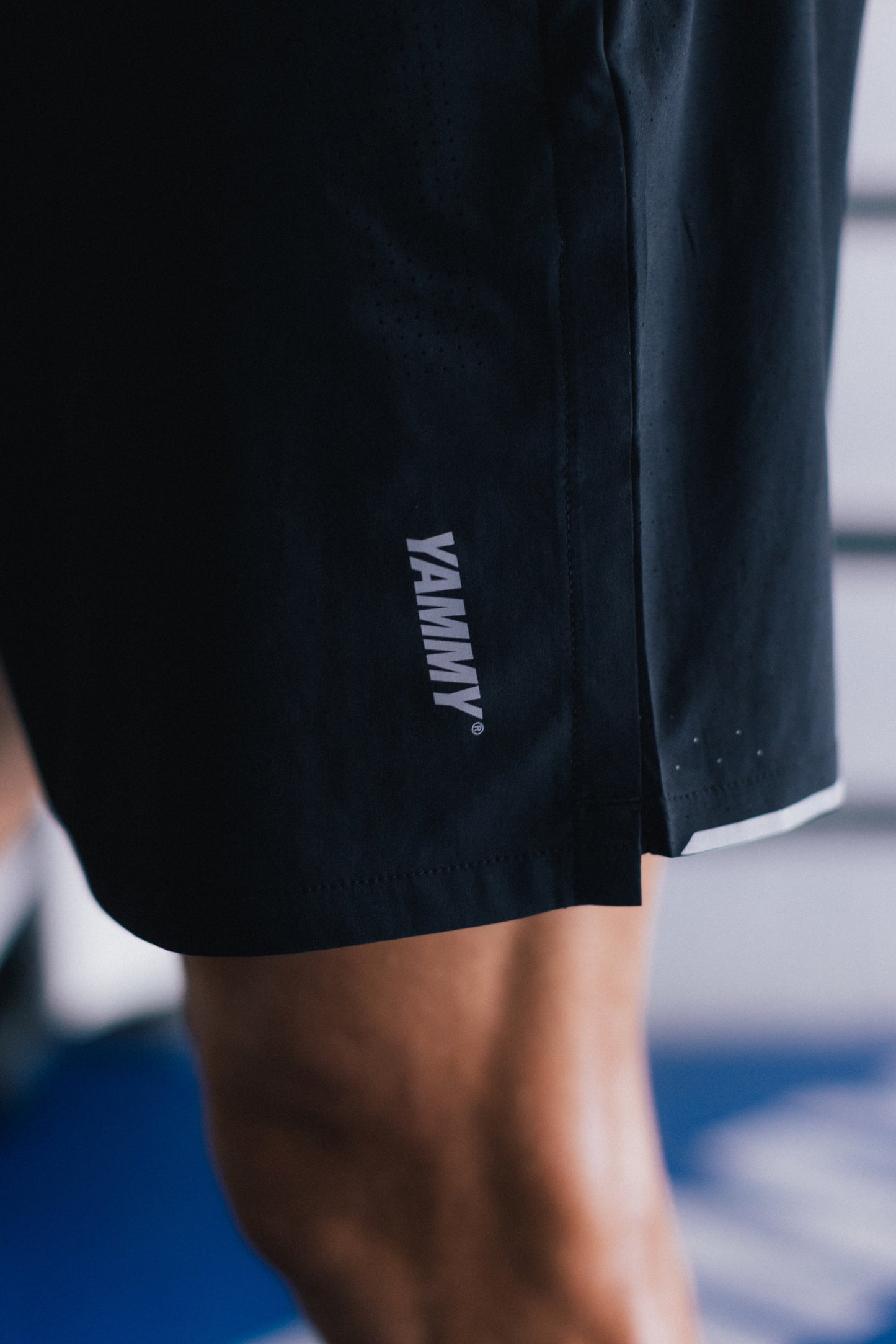Close-up of a person wearing black athletic shorts with a white vertical YAMMY logo on the lower left leg, photographed in a gym setting.