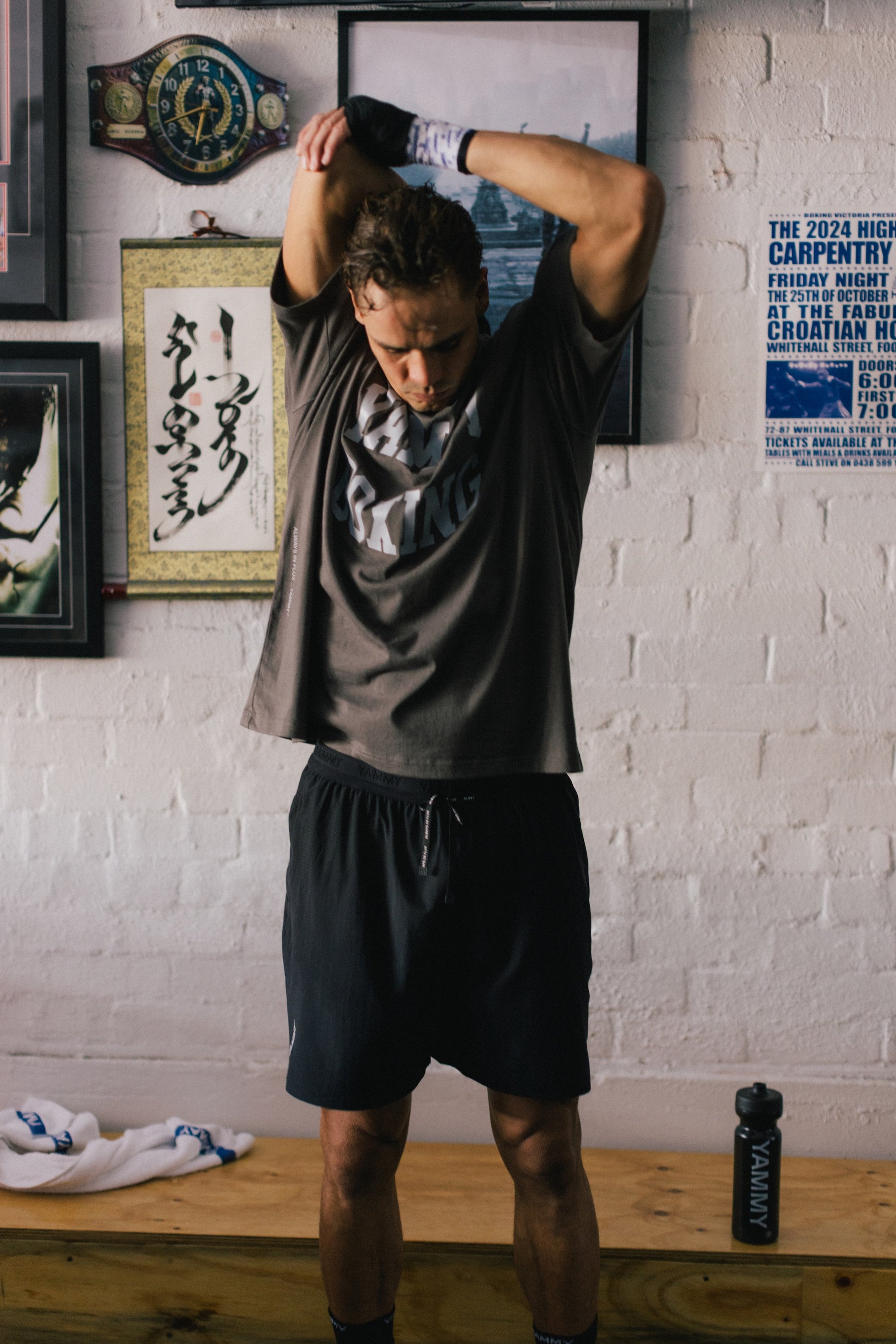 A man in a gym setting, wearing a dark gray t-shirt and black athletic shorts, stretching his arms above his head. The background features a white brick wall decorated with framed pictures, a championship belt, and a blue poster.