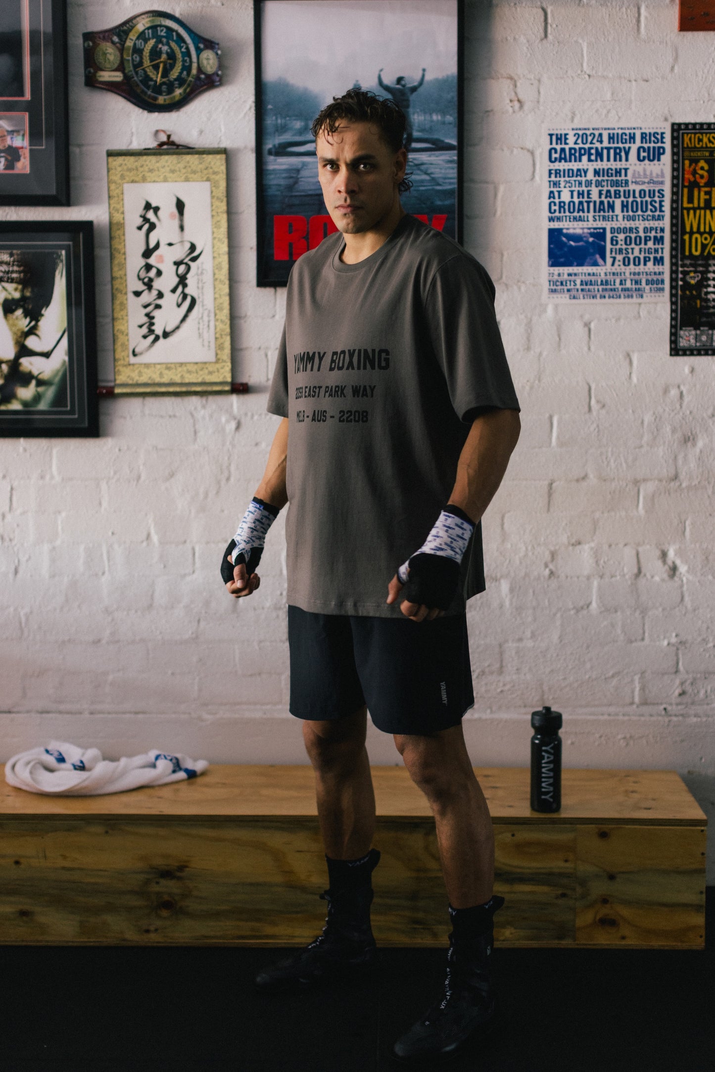 A man stands in a gym wearing a gray t-shirt with 'YAMMY BOXING' printed on the front, black boxing shorts, and black boxing boots. The background features a white brick wall with framed posters and a wooden bench.