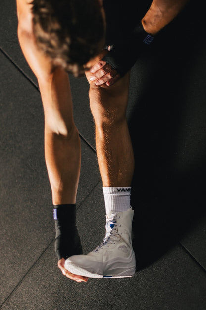 A close-up lifestyle shot of a person in a gym wearing a white high-top boxing shoe, white socks with 'yammy' printed on them, and black hand wraps.