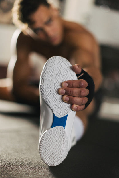 A close-up of a person stretching their leg while wearing a white athletic shoe with a blue accent and black heel wrap, set in a gym environment.