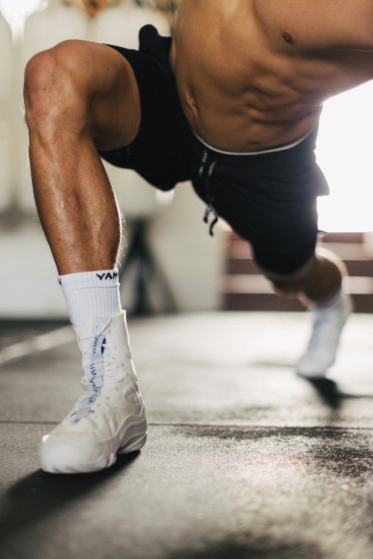 A close-up action shot of a man in a gym performing a lunge, wearing white athletic socks with blue branding and white sneakers, black athletic shorts, and a bare torso.