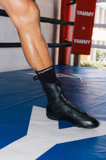 A close-up of a person's leg and foot wearing a black YAMMY brand boxing boot and matching black socks inside a boxing ring with blue and white flooring and red ropes.
