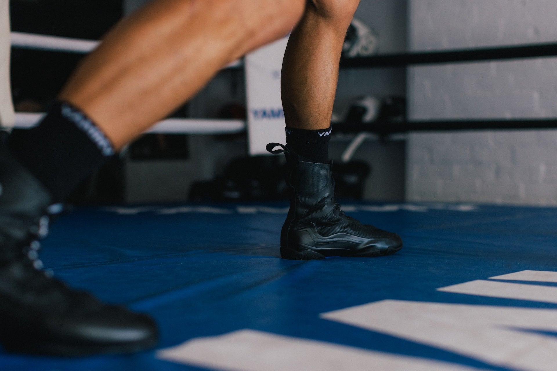 A close-up lifestyle shot of a boxer's legs and feet inside a boxing ring, wearing black leather boxing boots, black athletic socks, and black boxing shoes on a blue canvas floor.