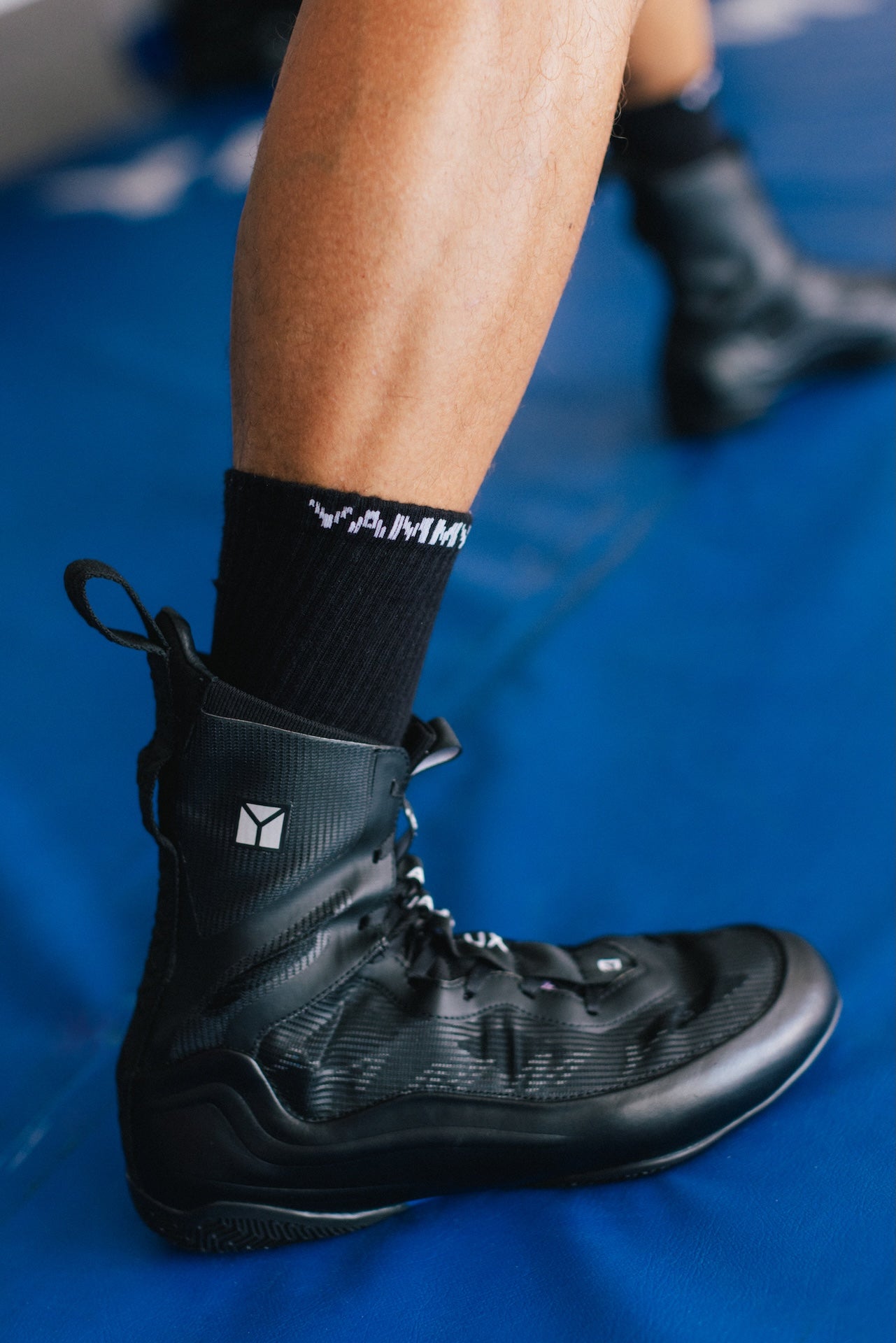 A close-up of a person's lower leg wearing a black Yammy athletic boot with black socks, standing on a blue mat.