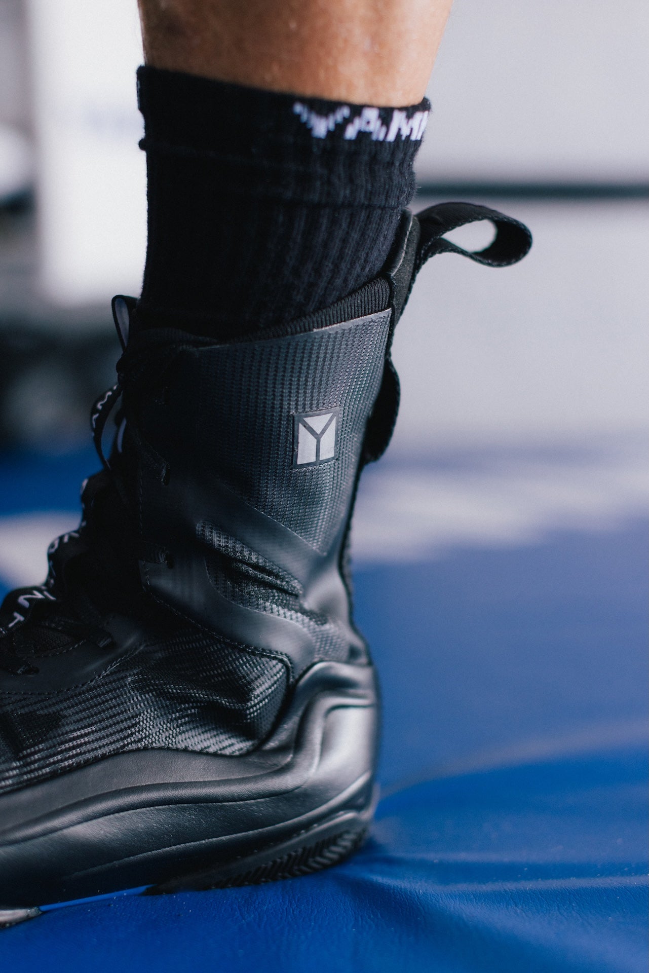 A close-up of a black athletic boot worn on a foot, paired with a black crew sock, set against a blue mat and blurred background.